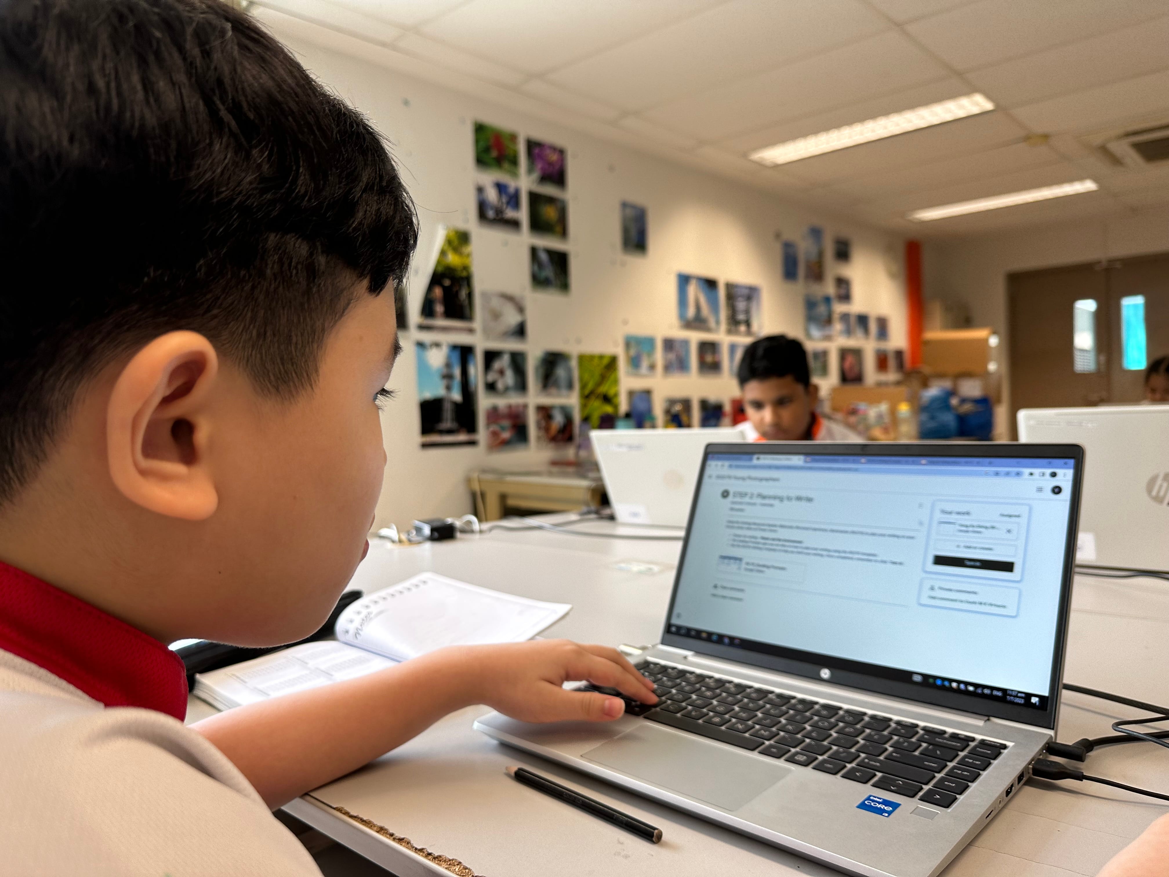 A young boy in a classroom works on an HP laptop displaying a writing assignment, with photos on the wall behind him.